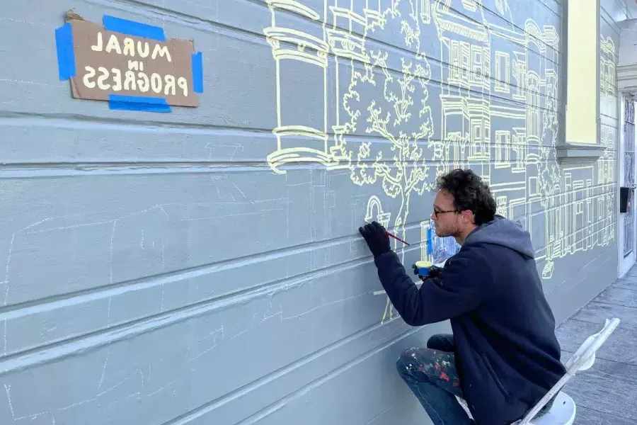 An artist paints a mural on the side of a building in 教会区, 在建筑上贴了一个牌子，上面写着“壁画正在进行中”.加州贝博体彩app.